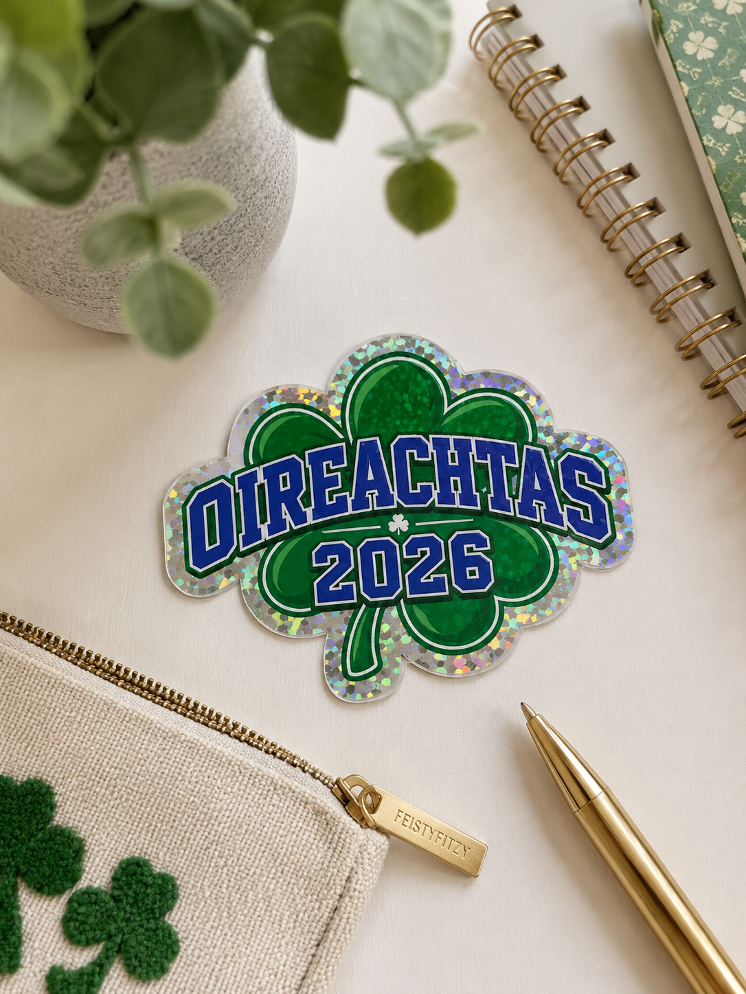 A St. Patrick's Day themed enamel pin shaped like a shamrock with the text 'OIREACHTAS 2026' in blue letters, displayed on a styled flat-lay desk with a green pencil pouch, gold pen, notebook, and plant.