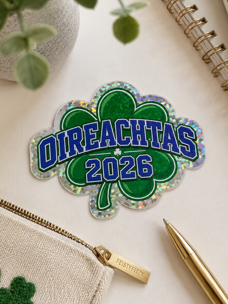 A St. Patrick's Day themed enamel pin shaped like a shamrock with the text 'OIREACHTAS 2026' in blue letters, displayed on a styled flat-lay desk with a green pencil pouch, gold pen, notebook, and plant.