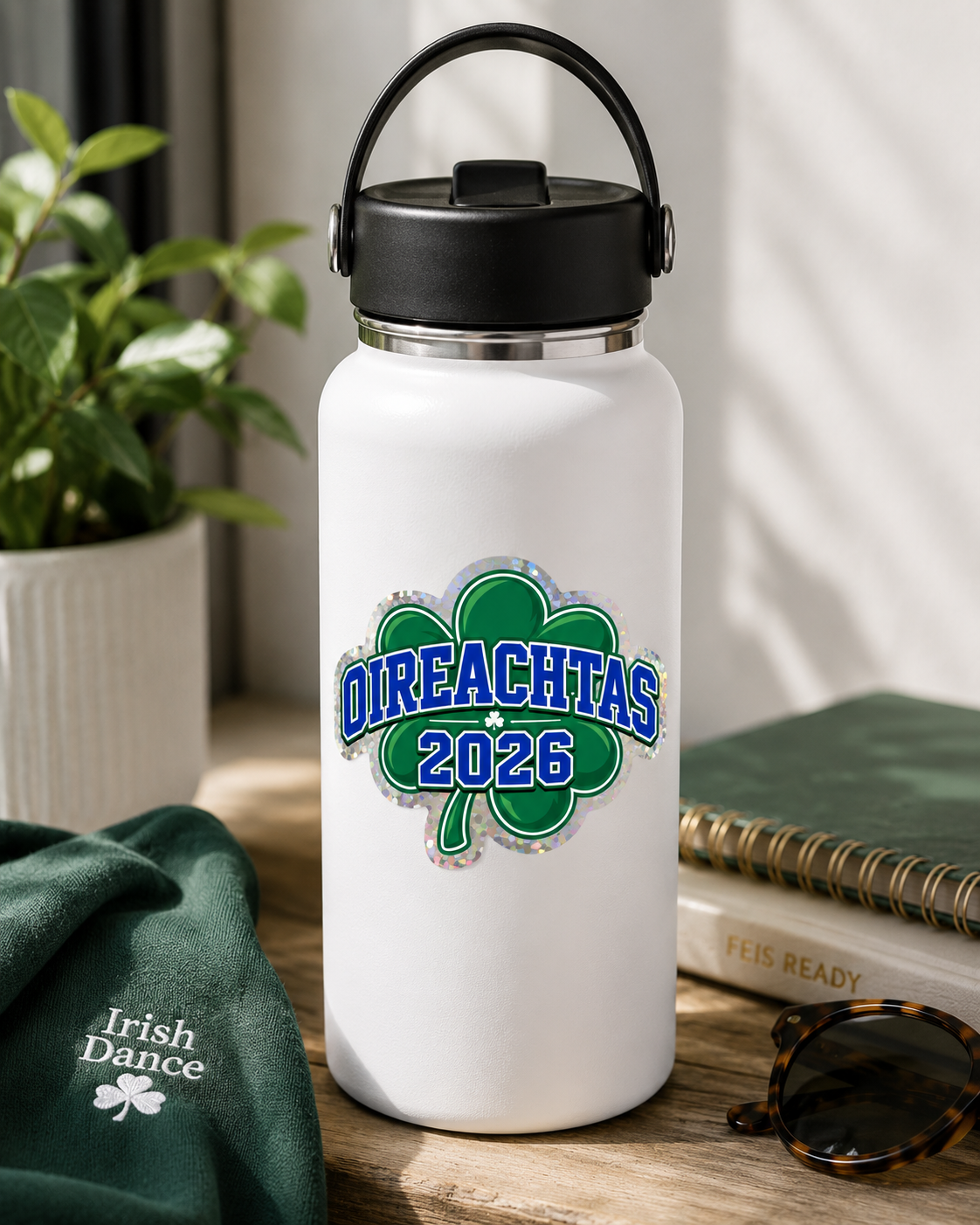 A white insulated water bottle with a black lid and carry handle, featuring a green shamrock graphic and the text 'DIREACHTAS 2026' on the front. The bottle is styled on a wooden table with a green 'Irish Dance' t-shirt, a notebook, sunglasses, and a potted plant in the background.