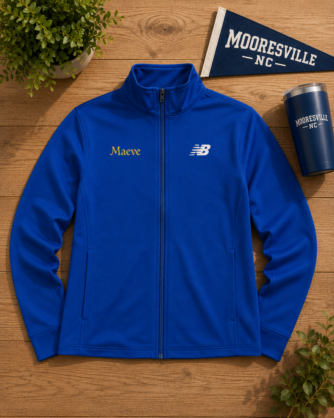 A royal blue zip-up jacket laid flat on a wooden surface, with a gold embroidered 'Maeve' name on the left chest and a white New Balance logo on the right chest. A navy 'Mooresville NC' flag, matching tumbler, and small plants are arranged around it as props.