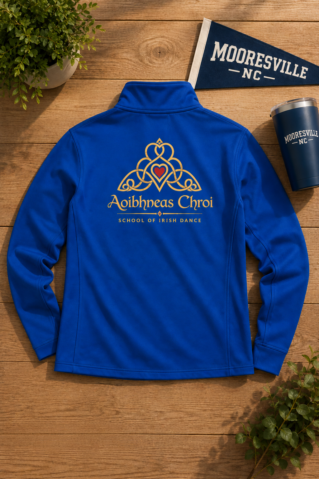 A royal blue quarter-zip athletic jacket is laid flat on a wooden surface, showing the back with a gold and red Celtic knot design and the text 'Aibhneas Chroi' and 'School of Irish Dance.' A small 'Mooresville NC' flag, matching cup, and potted plant are arranged around it as props.