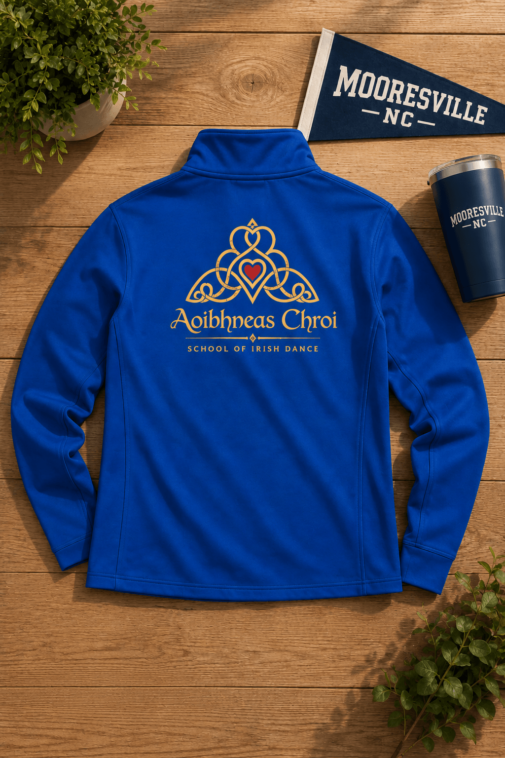 A royal blue quarter-zip athletic jacket is laid flat on a wooden surface, showing the back with a gold and red Celtic knot design and the text 'Aoibhneas Chroi' and 'School of Irish Dance.' A small 'Mooresville NC' flag, matching cup, and potted plant are arranged around it as props.
