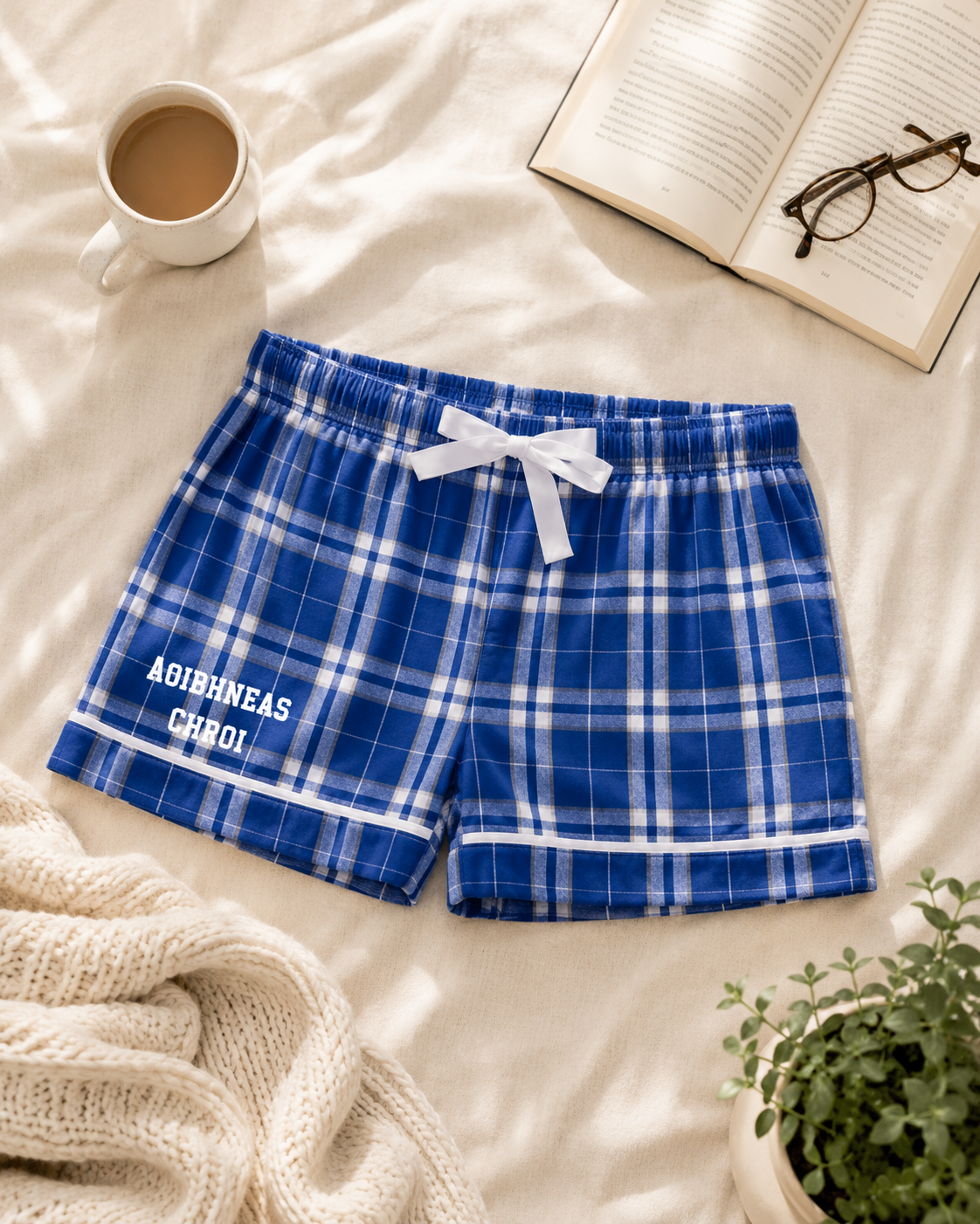 Blue and white plaid lounge shorts with a white drawstring and 'Aoibhineas Chroi' printed on the left leg, styled on a cream-colored bed with a mug, open book, glasses, knit blanket, and small plant.
