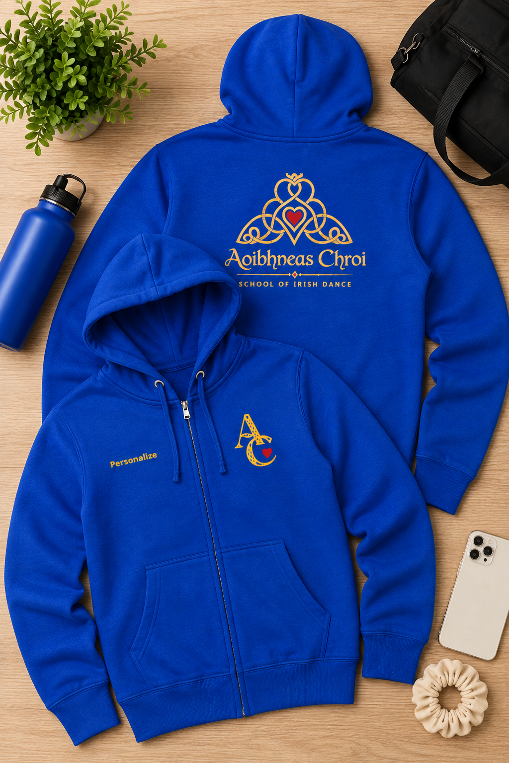 A royal blue zip-up hoodie and matching zip-up jacket laid flat on a light wood surface, styled with a water bottle, plant, black bag, phone, and scrunchie. The back of the hoodie shows a large gold Celtic knot and heart emblem with the text 'Aoibhneas Chroi' and 'School of Irish Dance.