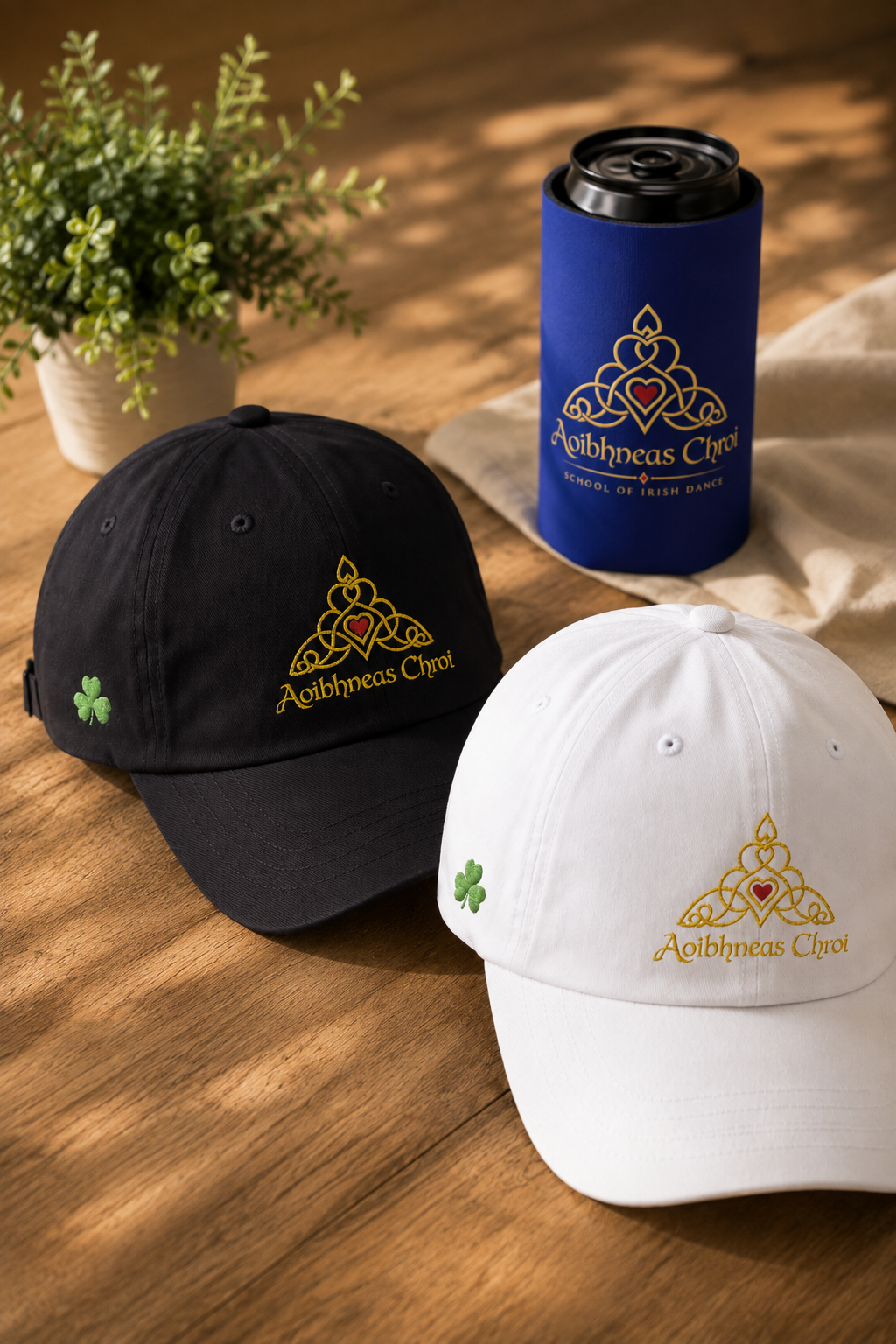 Two embroidered baseball caps, one black and one white, displayed on a wooden surface alongside a blue can cooler. The caps feature a gold Celtic knot logo with a red heart and the text 'Aoibhneas Chroi ', along with a small green shamrock on the side.