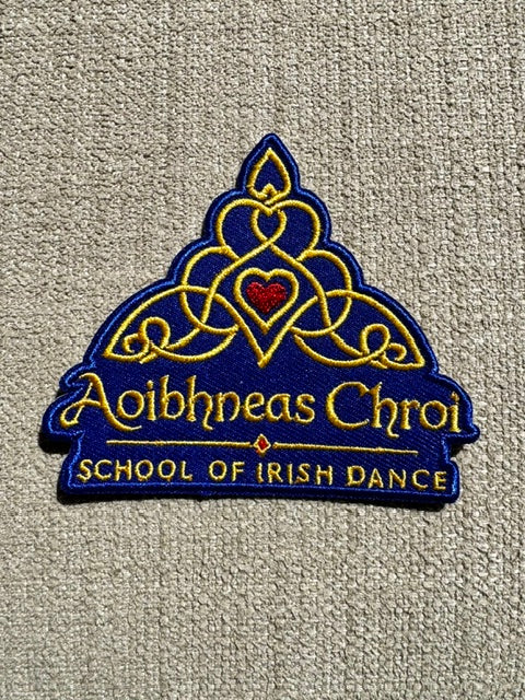 An embroidered patch for the Aoibhneas Chroi School of Irish Dance, featuring a blue and gold Celtic knot design with a small red heart in the center, displayed on a beige textured fabric background.