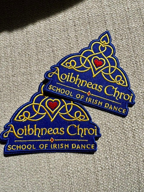 Two embroidered patches for 'Aoibhneas Chroi - School of Irish Dance' in blue and gold, displayed on a textured beige carpet.