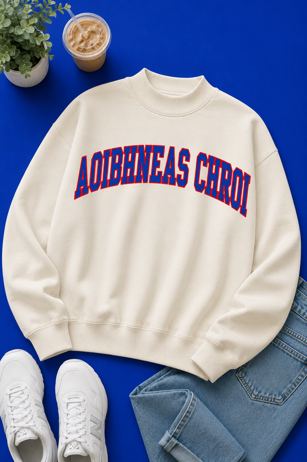 An off-white crewneck sweatshirt with 'AOIBHNEAS CHROI' printed in red and blue block letters across the chest, styled in a flat lay with white sneakers, blue jeans, an iced coffee, and a small potted plant on a vibrant blue background.