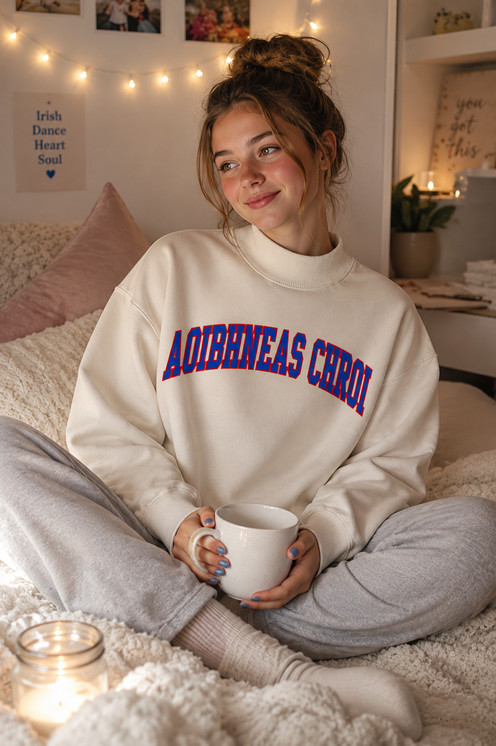 A woman sitting cross-legged on a cozy bed in a warmly lit bedroom, wearing a cream sweatshirt with 'AOIBHEAS CHROI' in blue and red lettering, holding a white mug.