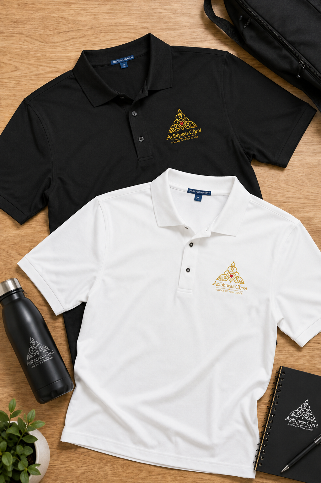 Two polo shirts, one black and one white, laid flat on a light wood surface with a small gold embroidered logo on the left chest. A black water bottle, notebook, and plant are placed around them as props.