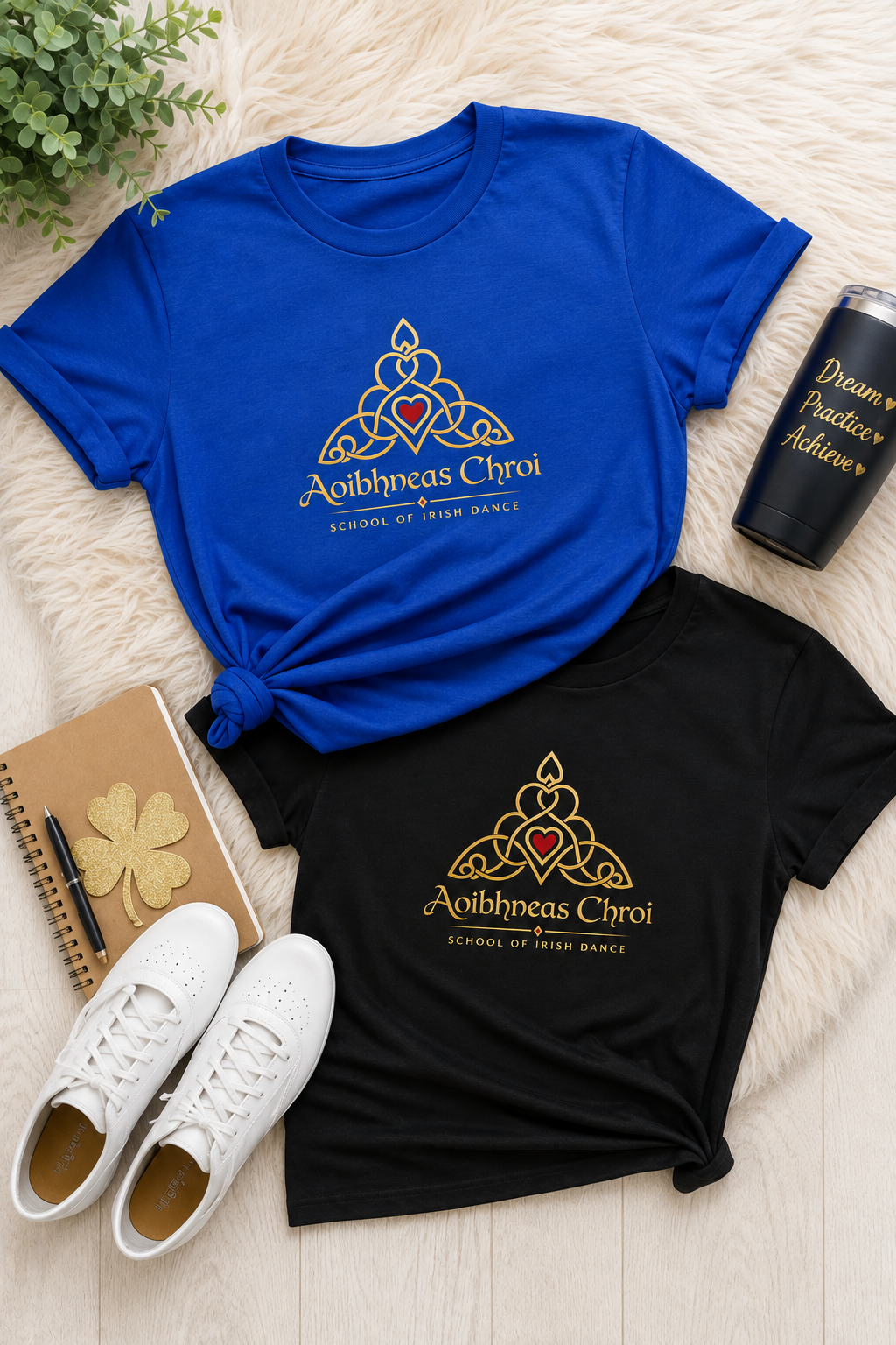 Two graphic t-shirts, one royal blue and one black, laid flat on a white faux fur surface with lifestyle props including white sneakers, a notebook, and a black can.