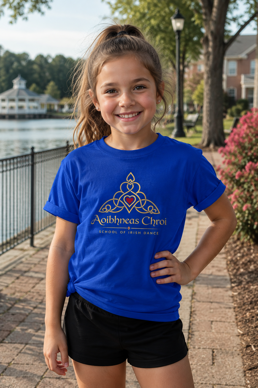A young girl smiling outdoors on a paved walkway near water, wearing a bright royal blue graphic T-shirt with a gold Celtic knot design and the text 'Aibhneas Chroi' and 'School of Irish Dance'.