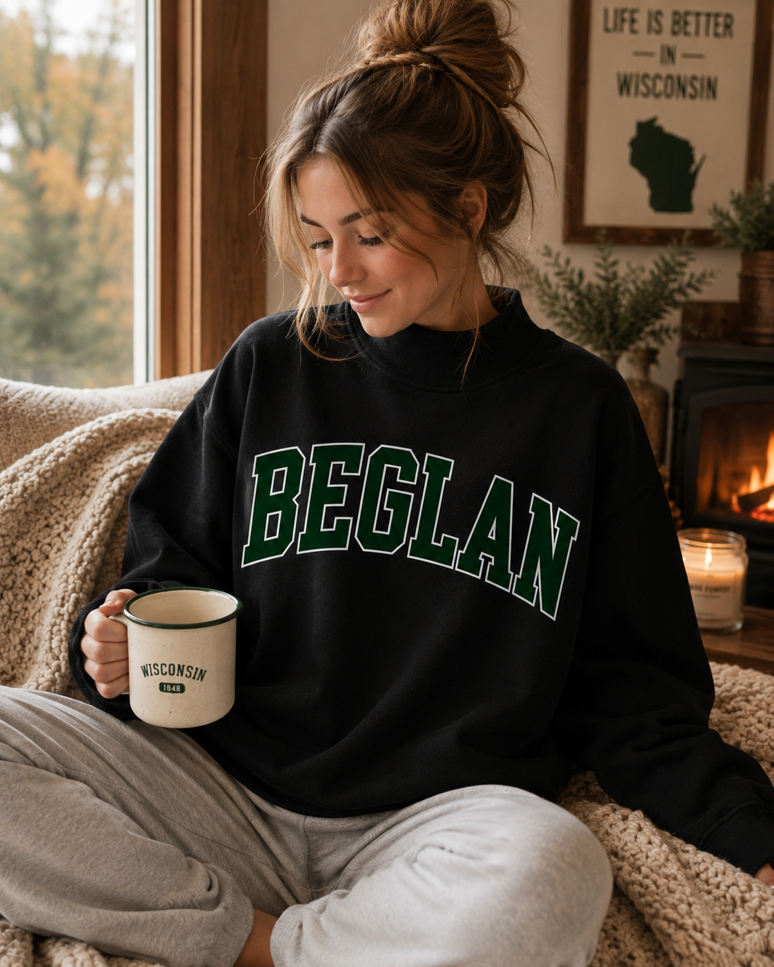 A woman sits on a cozy couch by a window, wearing a black sweatshirt with 'BEGLAN' in green varsity letters outlined in white. She holds a cream mug with 'WISCONSIN' printed on it.