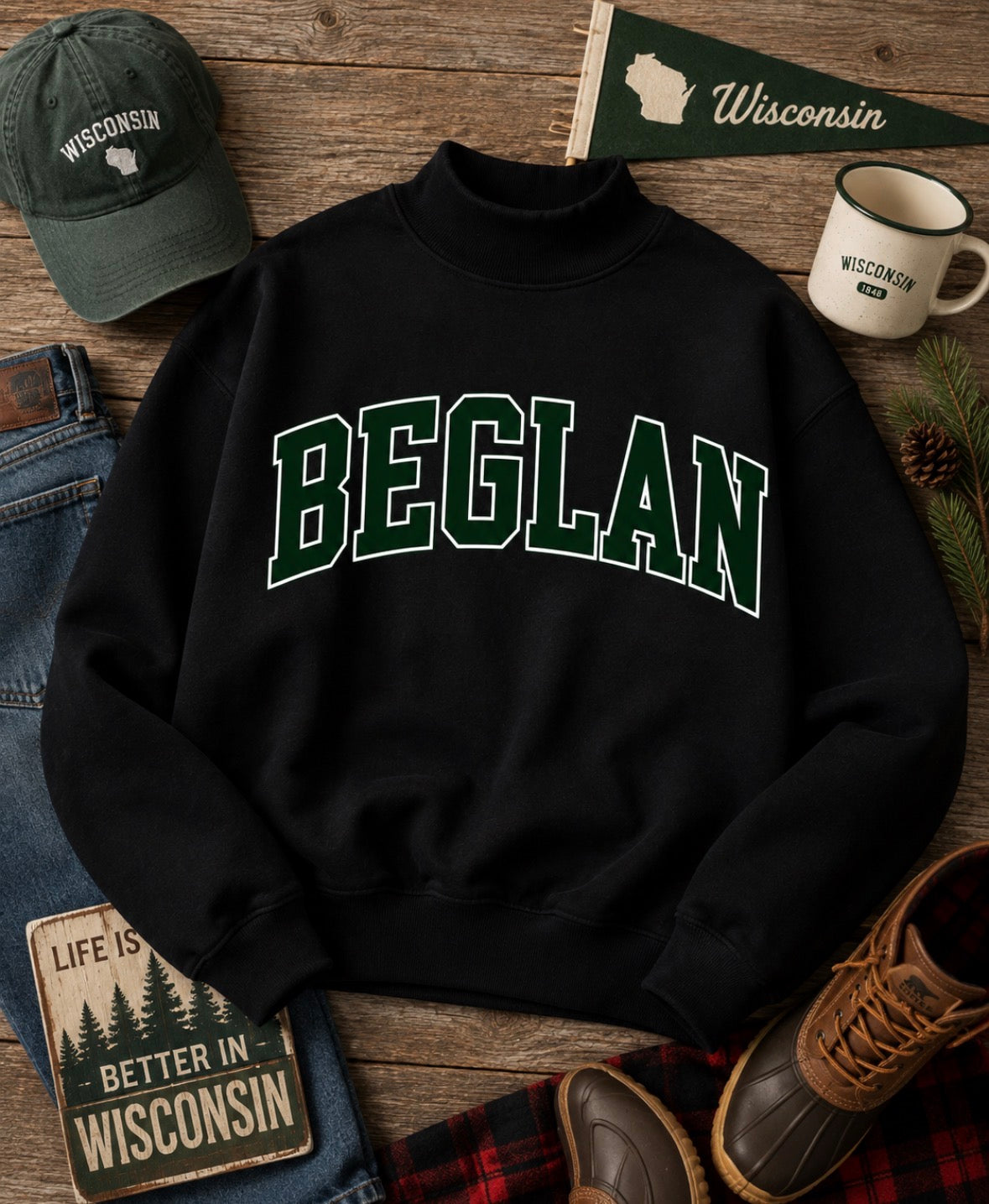 A black mock-neck sweatshirt with 'BEGLAN' printed in green varsity letters across the chest, styled in a rustic flat-lay with a green 'Wisconsin' cap, denim, a mug, boots, and a plaid scarf on a wooden surface.
