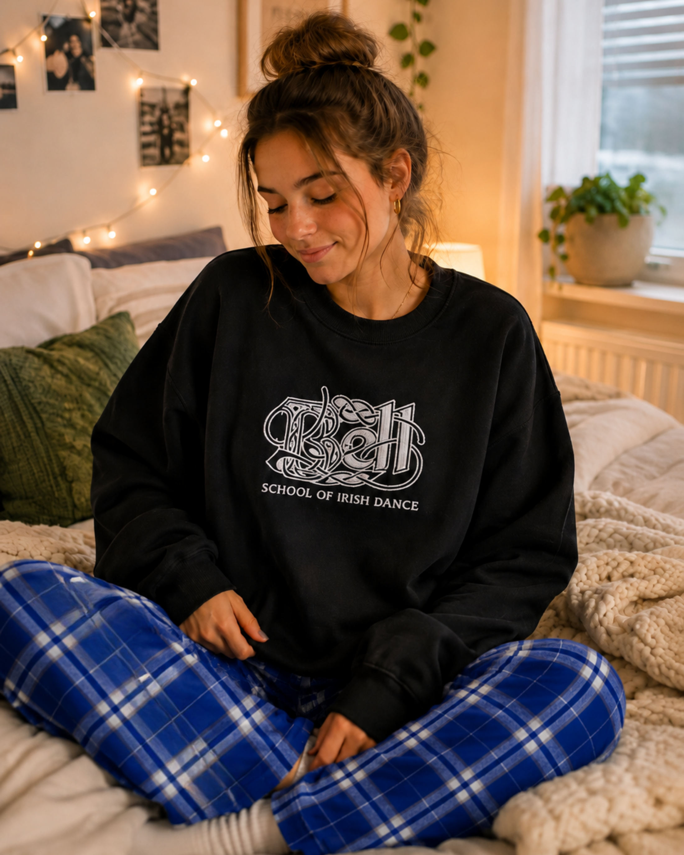 A woman sitting cross-legged on a bed wearing a black crewneck sweatshirt with a white graphic that reads 'School of Irish Dance' and blue plaid pants in a cozy bedroom setting.