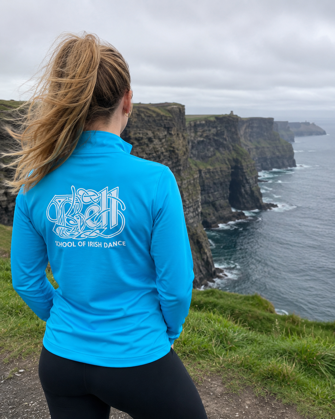 A woman seen from behind wearing a bright blue long-sleeve athletic top with a white graphic and the text 'SCHOOL OF IRISH DANCE' on the back, standing on a grassy cliffside overlooking the Cliffs of Moher.