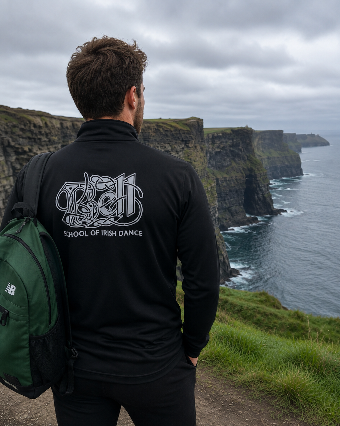 A man viewed from behind wearing a black long-sleeve quarter-zip sweatshirt with a white graphic print of the 'School of Irish Dance' on the back, standing on a grassy cliffside overlooking the Cliffs of Moher in Ireland. He is wearing a green New Balance backpack.