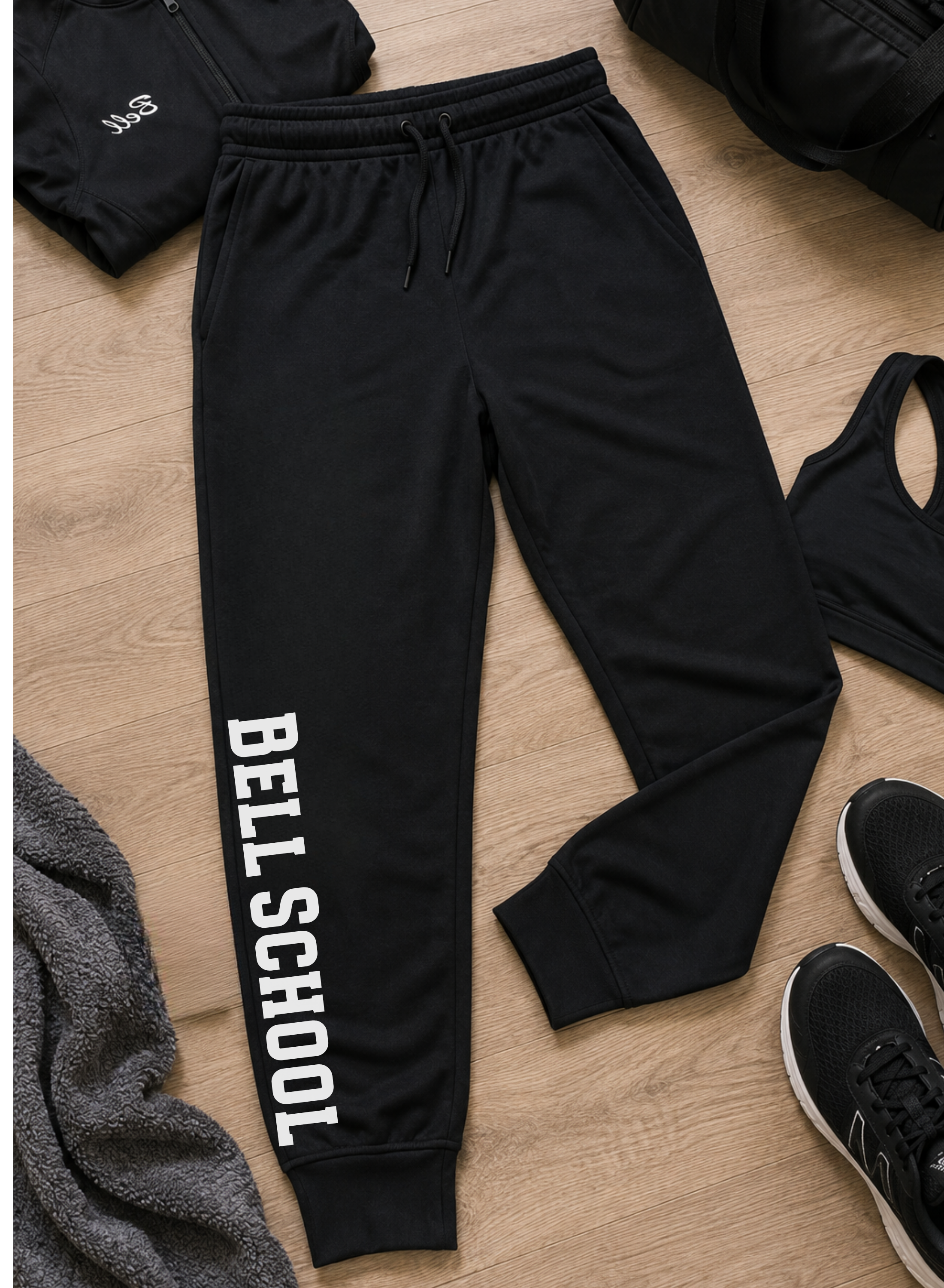 Black jogger sweatpants with 'BELL SCHOOL' printed vertically in white on the lower left leg, laid flat on a light wood floor with other athletic apparel and shoes around them.