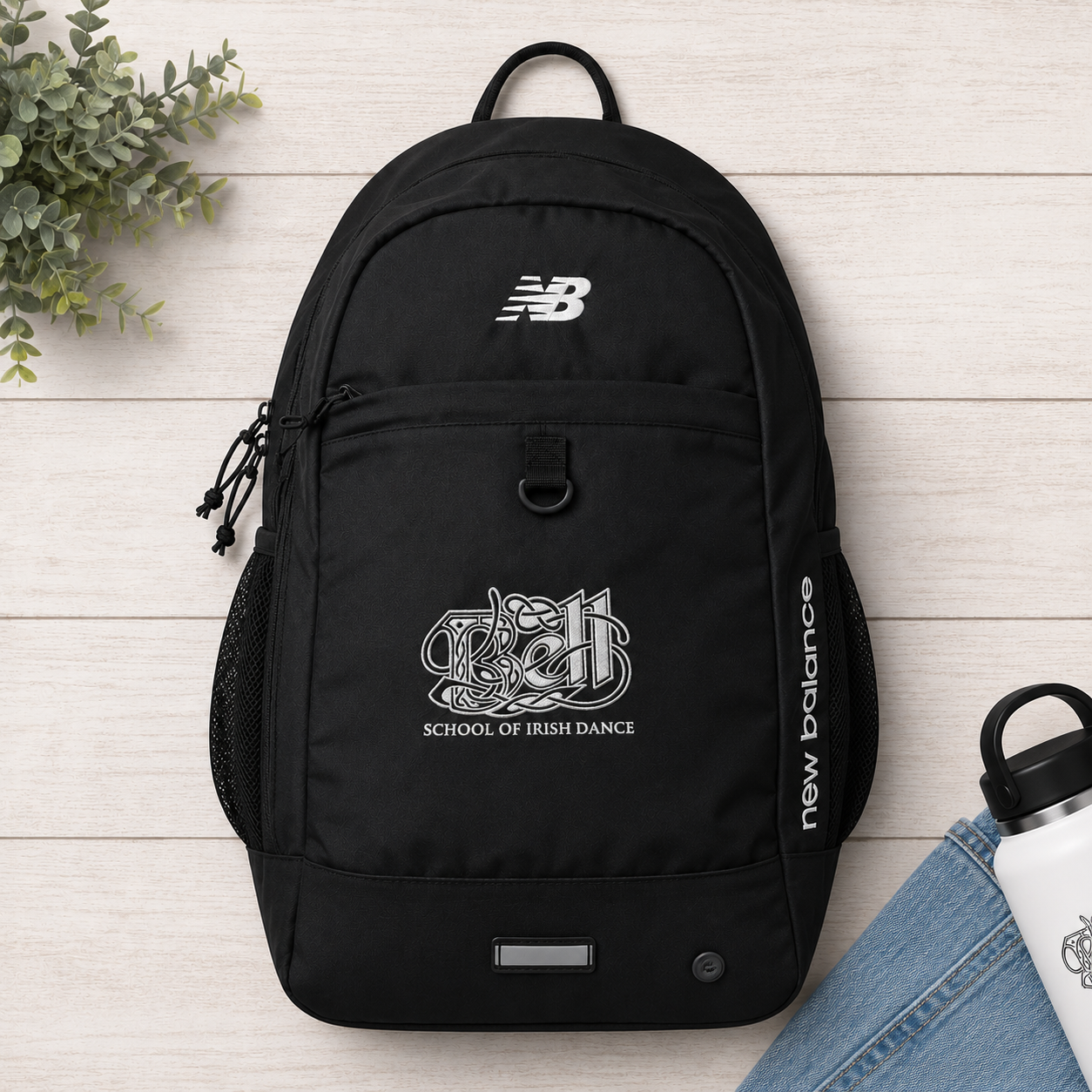 A black New Balance backpack with white printed logos, including 'School of Irish Dance' on the front pocket, displayed on a light wood background with a plant and denim props.