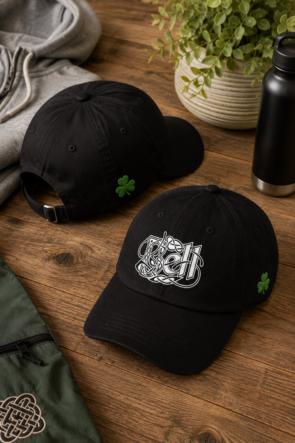 Two black baseball caps on a wooden tabletop. The front cap features a white embroidered Celtic knot logo and a small green shamrock on the side, while the rear cap shows the back view with a black buckle strap and a shamrock on the side.