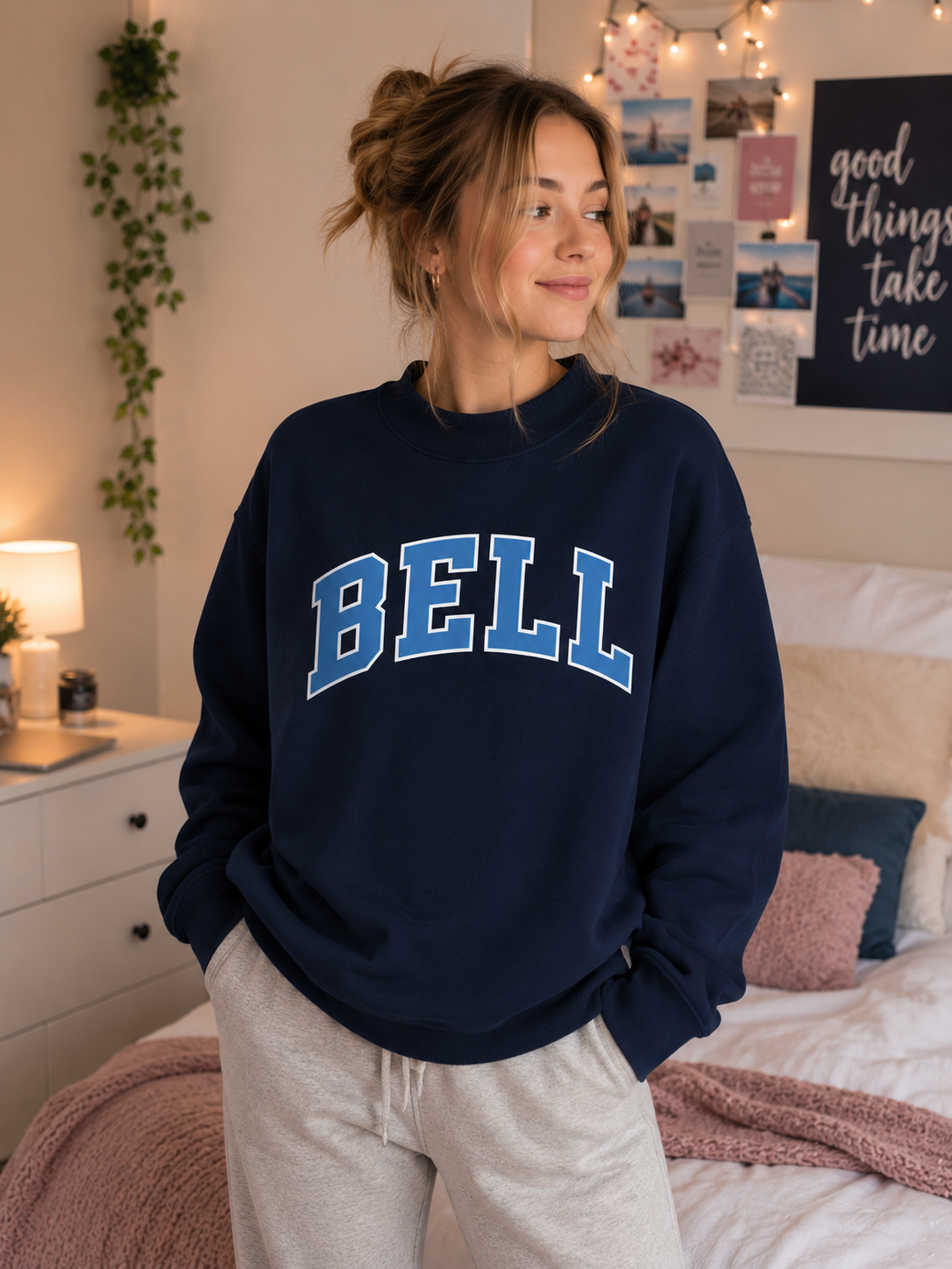 A woman wearing a navy blue crewneck sweatshirt with 'BELL' printed in blue varsity letters across the chest, paired with light gray sweatpants, standing in a cozy bedroom setting.