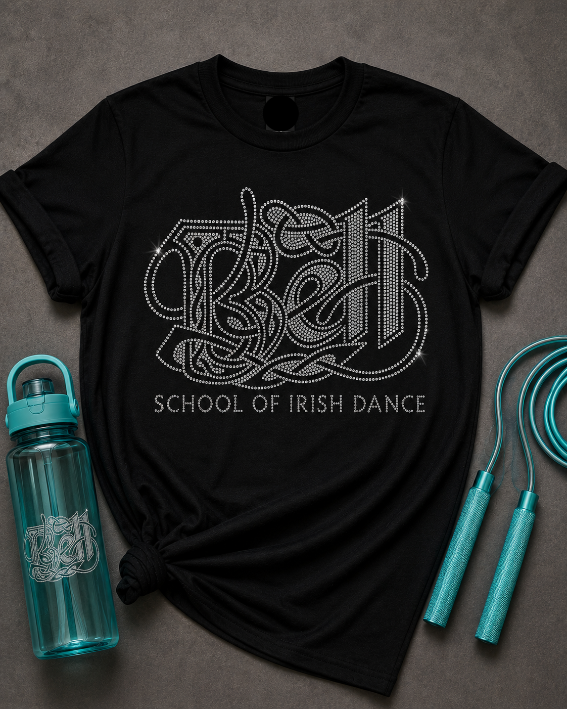 A black short-sleeve graphic T-shirt laid flat on a dark gray surface, styled with a turquoise water bottle, jump rope, and knotted hem. The shirt features a large rhinestone-style 'BEE' graphic with 'SCHOOL OF IRISH DANCE' text below.