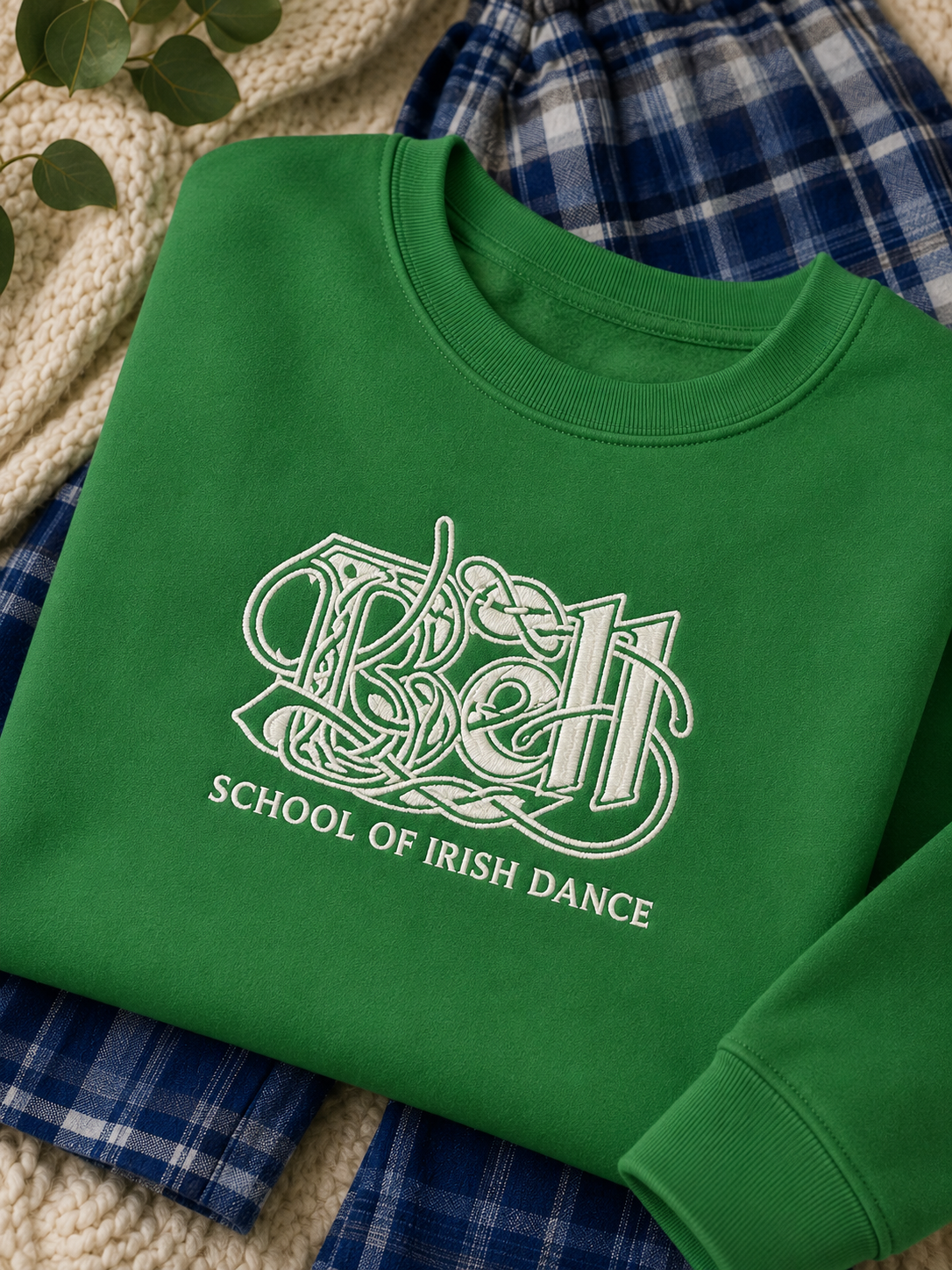 A green crewneck sweatshirt with a white graphic reading 'Belt' in stylized lettering and 'SCHOOL OF IRISH DANCE' underneath, laid flat on a cream knit blanket with blue plaid fabric and green leaves around it.