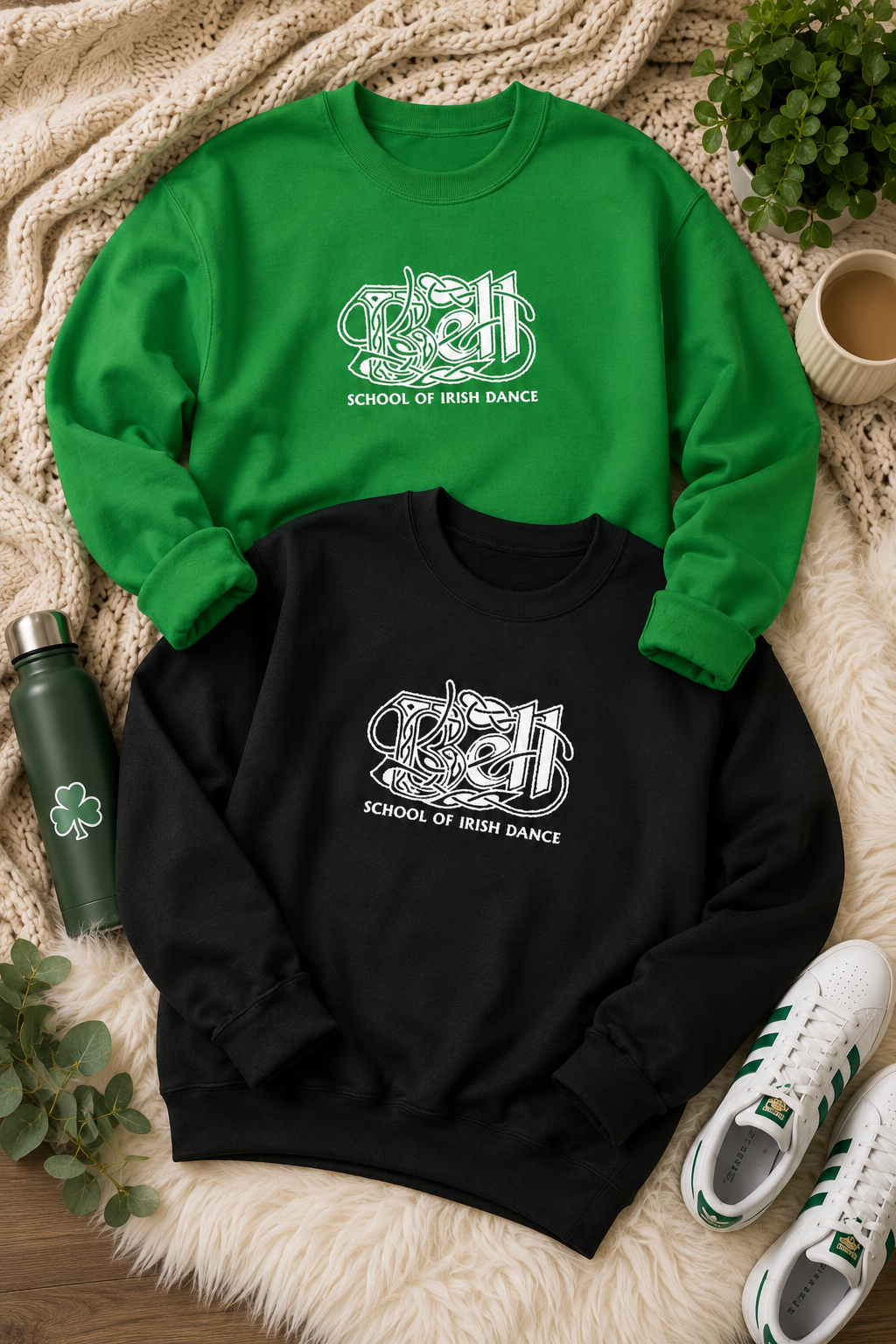 Two crewneck sweatshirts laid flat on a white faux-fur surface: a bright green one and a black one, both with a white Celtic knot graphic and the text 'SCHOOL OF IRISH DANCE' on the chest. Surrounding props include a green water bottle, white sneakers, a plant, and a cup on a wood surface.