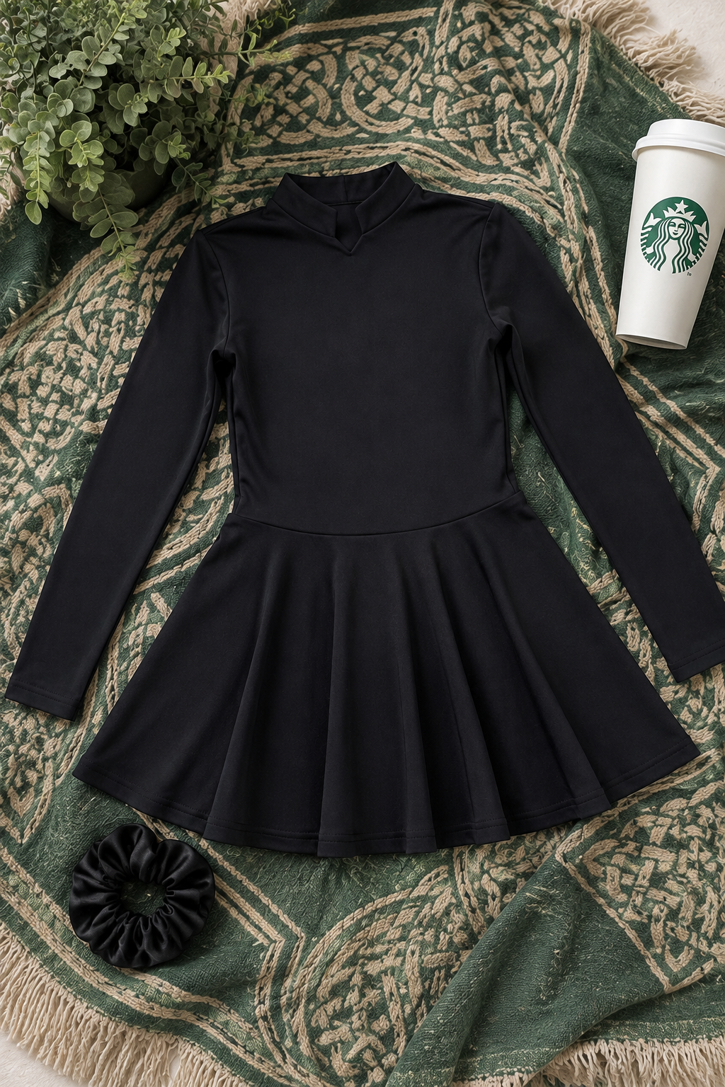 A black long-sleeve mock-neck dress with a flared skirt, laid flat on a green and beige patterned throw blanket. A Starbucks cup, a black scrunchie, and green foliage are styled around it.
