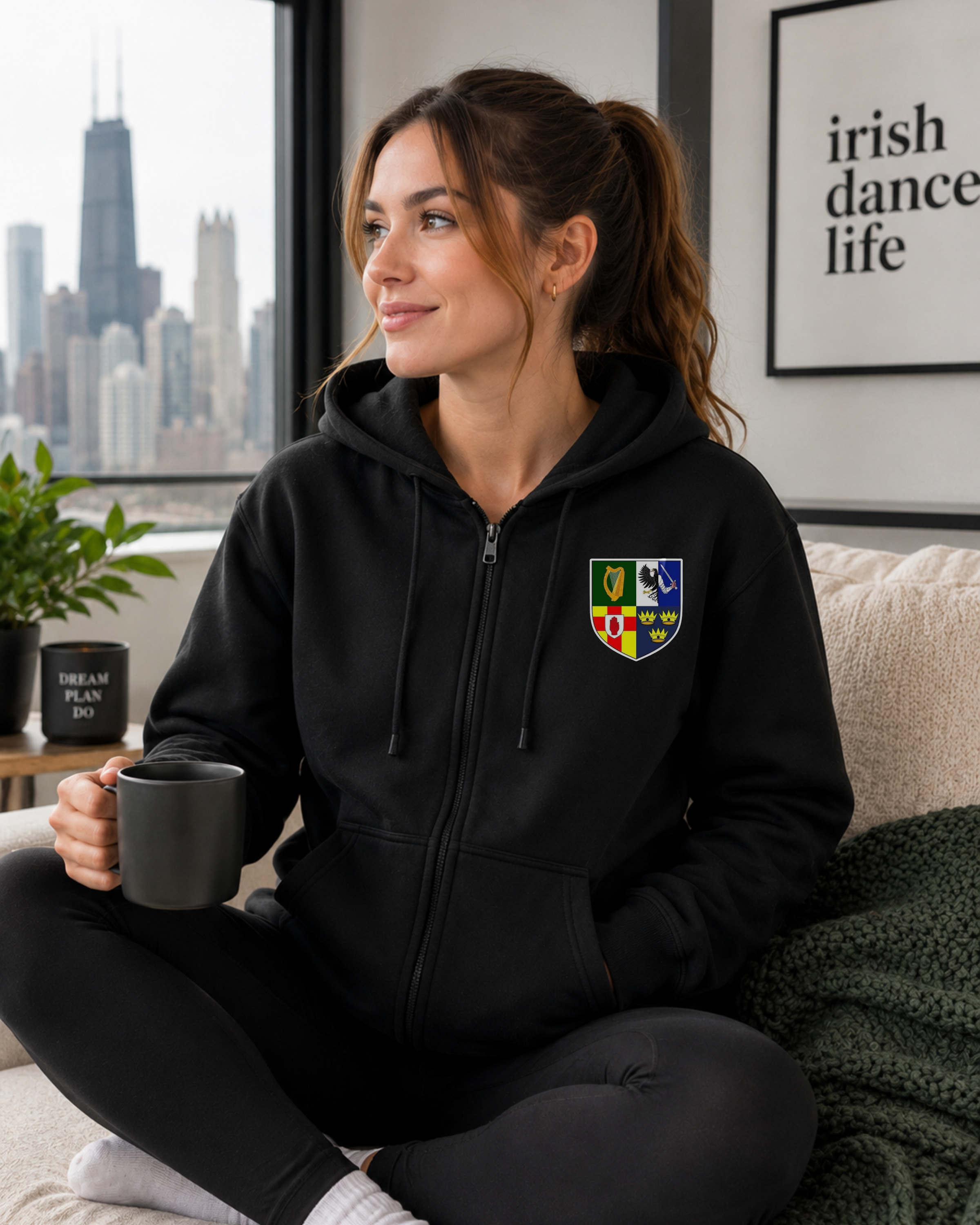 A woman sits cross-legged on a couch in a living room, wearing a black zip-up hoodie with a colorful shield crest on the left chest. She holds a dark gray mug, with a city skyline visible through the window behind her.