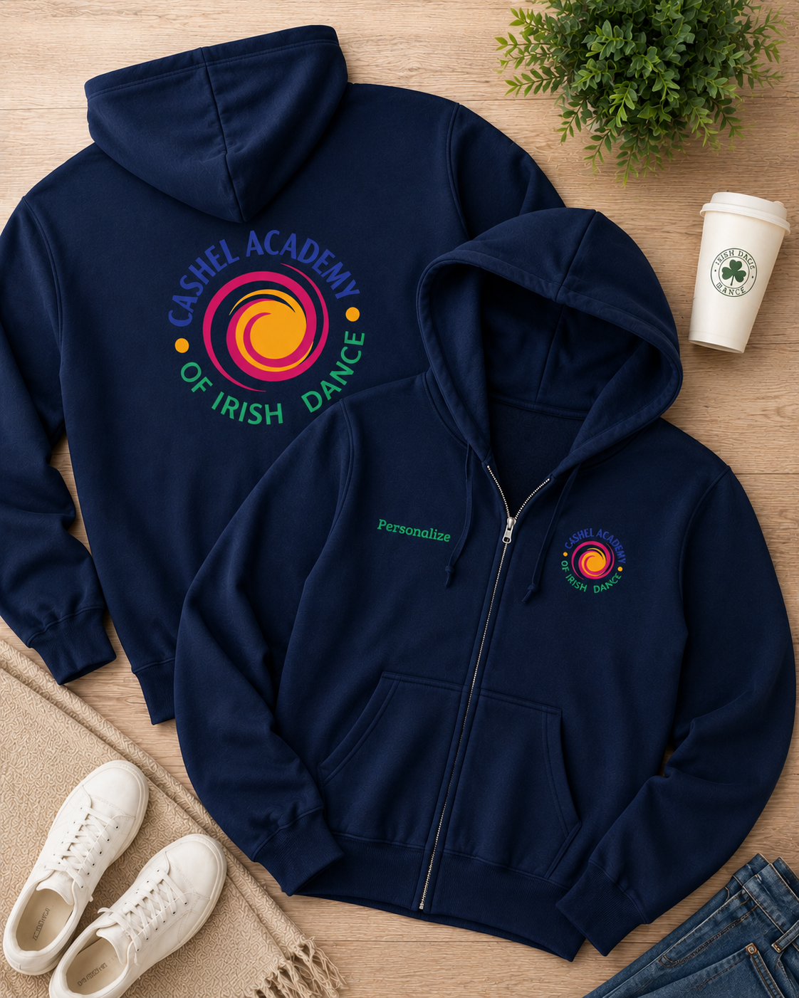 Two navy blue zip-up hoodies laid flat on a light wood surface, one showing the back with a large colorful circular logo and the text 'Cashel Academy of Irish Dance,' and the other showing the front with a small matching chest logo. White sneakers, a coffee cup, and jeans are placed around them as styling props.