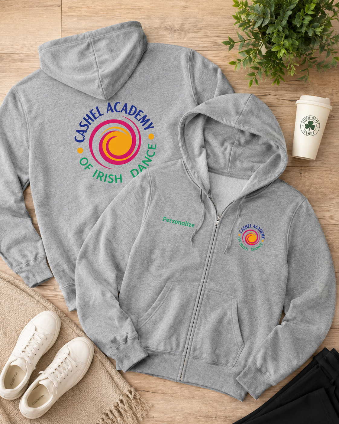 Two heather gray hoodies are arranged on a light wood floor with lifestyle props: a hoodie showing the back graphic and a zip-up hoodie showing the front chest logo. The graphics read 'Cashel Academy of Irish Dance' with a colorful spiral emblem.