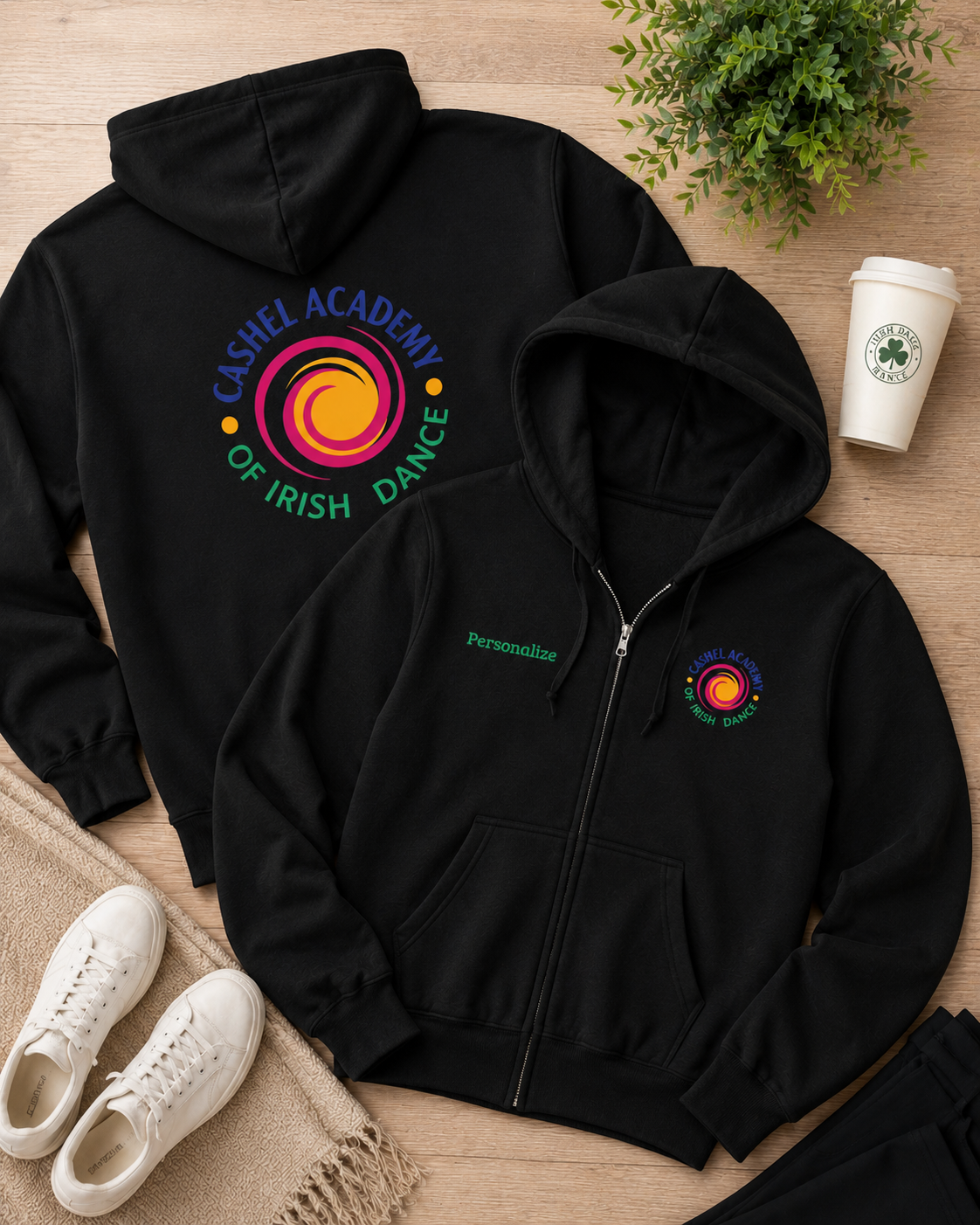 A black zip-up hoodie shown from both front and back on a wooden surface, styled with white sneakers, a plant, and a coffee cup. The front has a small multicolor circular logo and green 'Personalise' text, while the back has a large multicolor circular logo reading 'Cashel Academy of Irish Dance'.