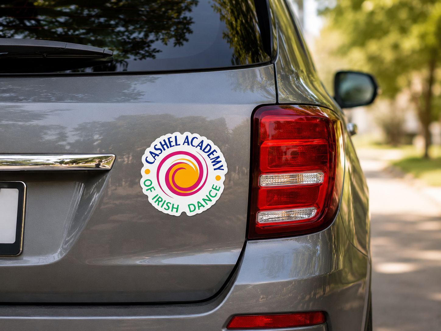 A circular sticker for 'Cashel Academy of Irish Dance' applied to the rear left side of a gray vehicle, with a blurred outdoor background showing trees and a path.