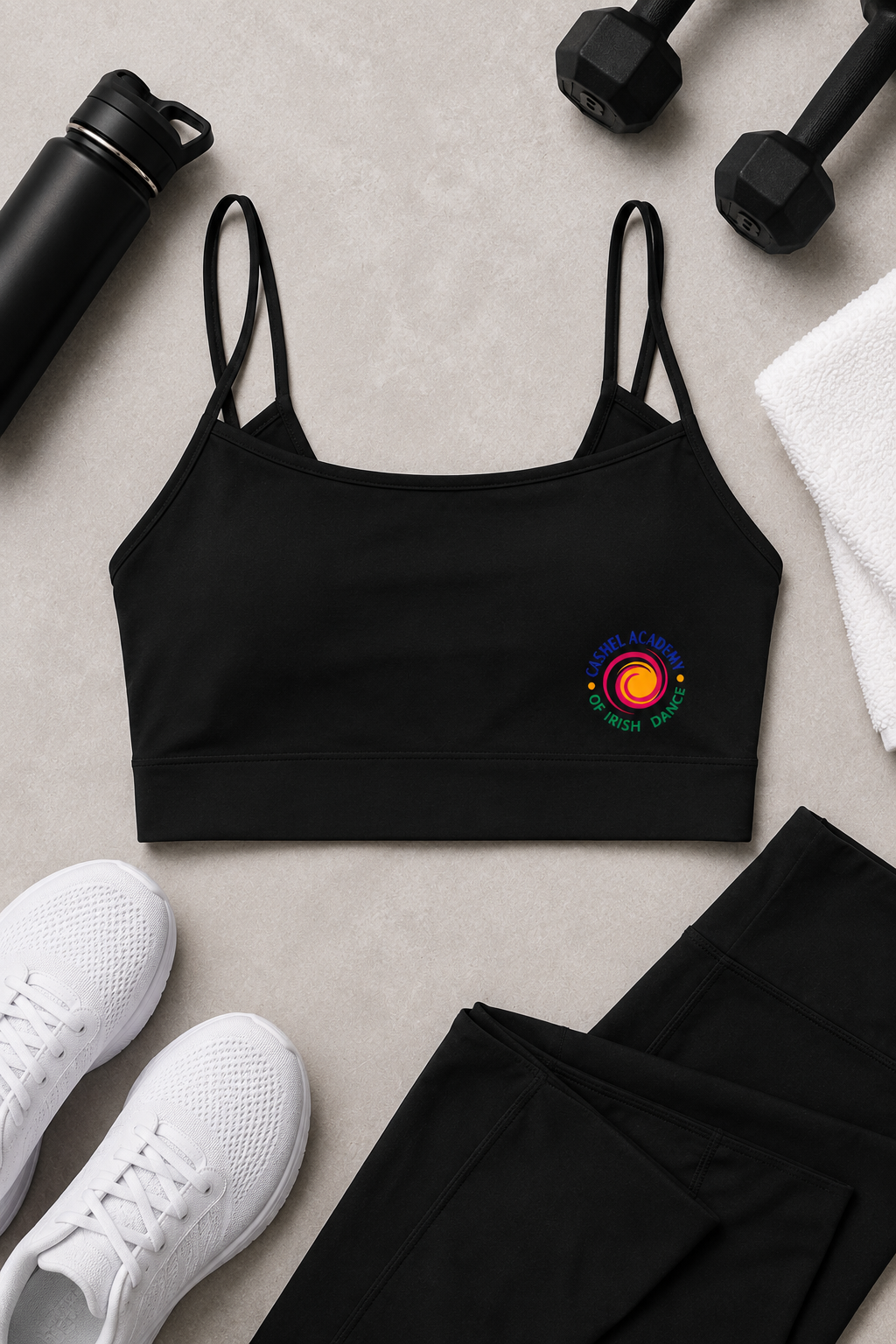 A black sports bra with thin straps and a colorful circular graphic on the left chest, styled in a flat lay with black leggings, white sneakers, a black water bottle, dumbbells, and a white towel on a light gray surface.