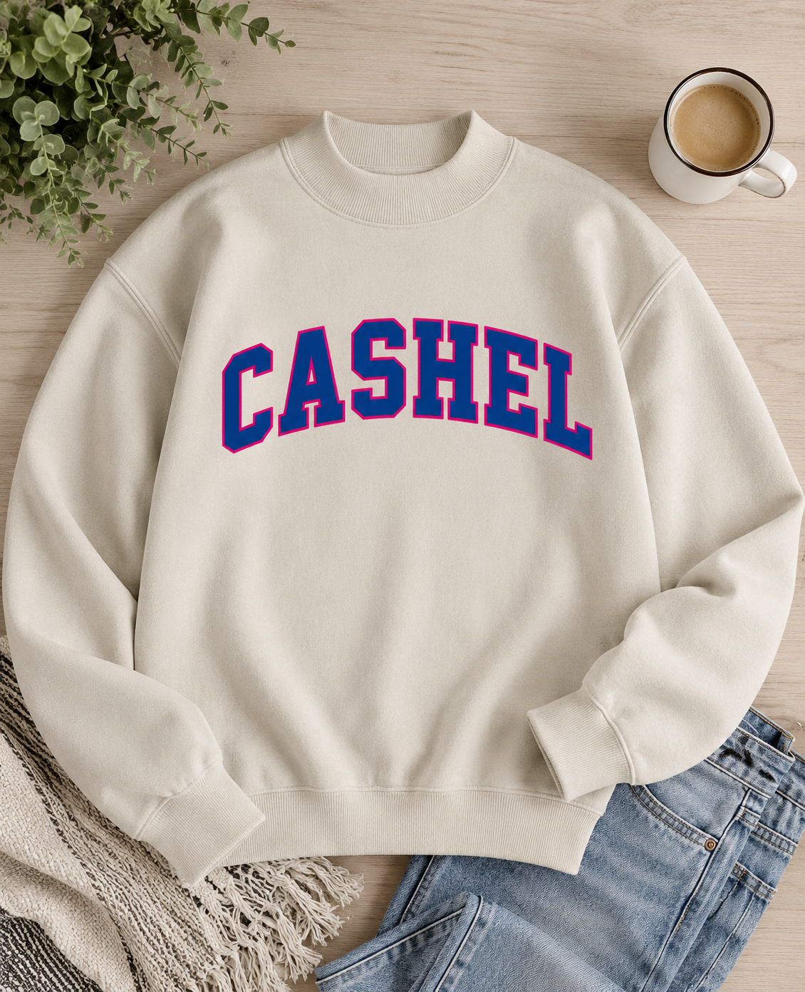 An off-white crewneck sweatshirt with 'CASHEL' embroidered in blue and pink block letters across the chest, styled in a flat lay with jeans, a coffee cup, and a plant on a light wood background.