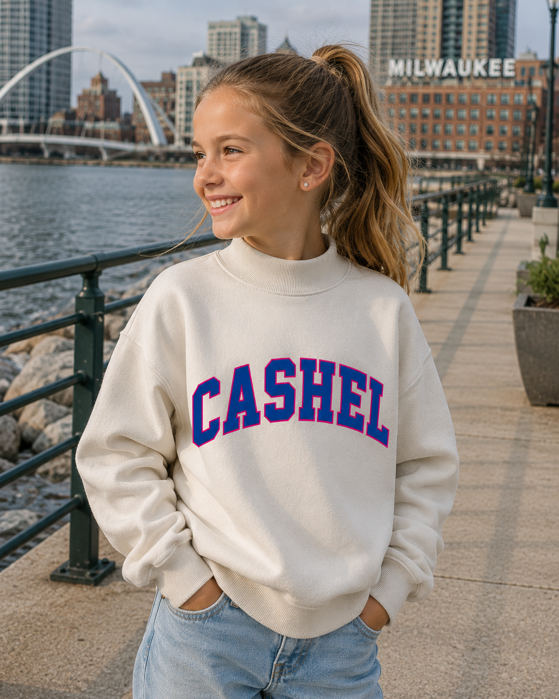 A young girl with her hair in a ponytail stands on an outdoor waterfront walkway wearing a cream-colored crewneck sweatshirt with 'CASHEL' printed in blue and red varsity lettering across the chest. The background shows a city skyline with the Milwaukee sign and a bridge.
