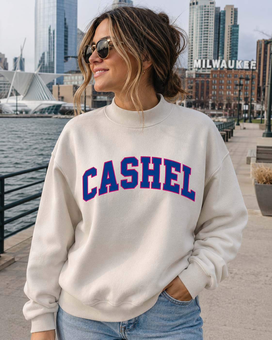 A woman wearing a cream-colored crewneck sweatshirt with 'CASHEL' printed in blue and red varsity lettering across the chest, standing outdoors near a waterfront with a city skyline in the background.
