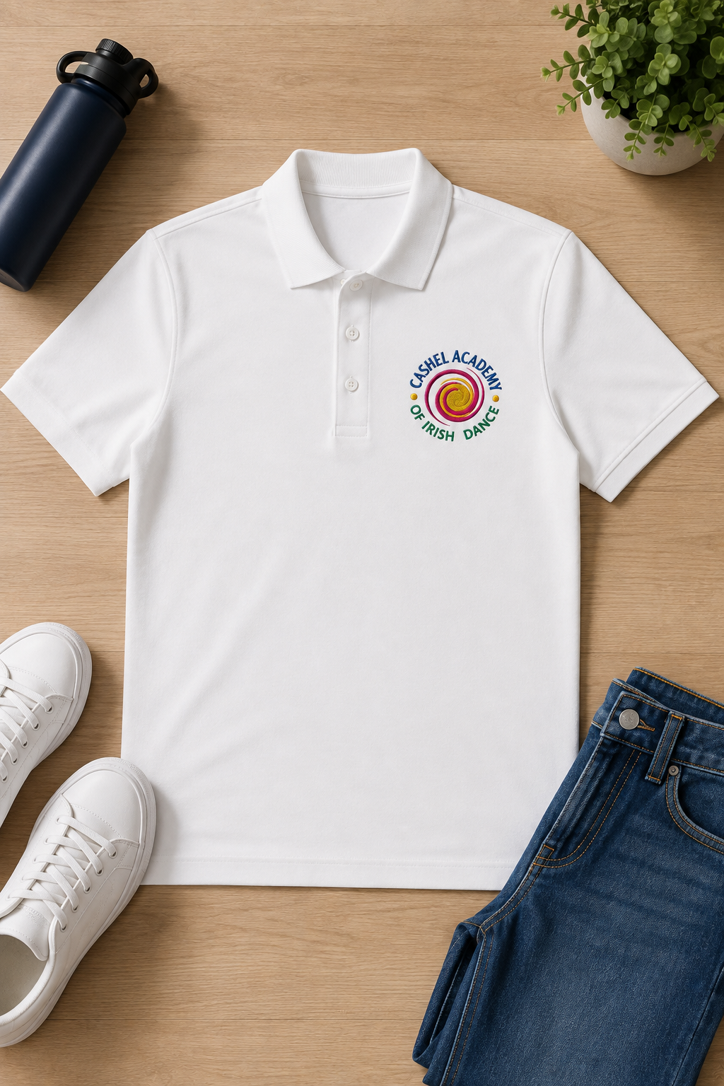 A white short-sleeve polo shirt laid flat on a light wood surface, featuring a colorful circular chest logo that reads 'Cashel Academy of Irish Dance.' White sneakers, blue jeans, a dark bottle, and a small potted plant are arranged around it as styling props.