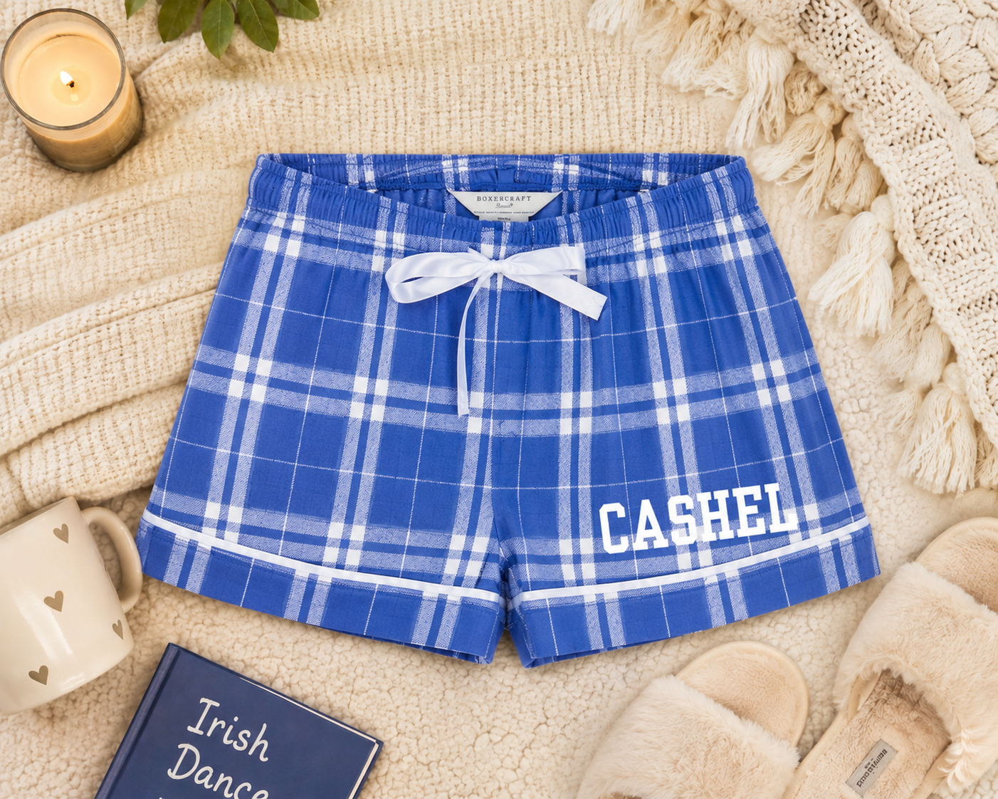 Blue and white plaid lounge shorts with a white drawstring and 'CASHEL' printed in white on the left leg, styled in a cozy flat-lay with a candle, mug, book, slippers, and knit blanket.