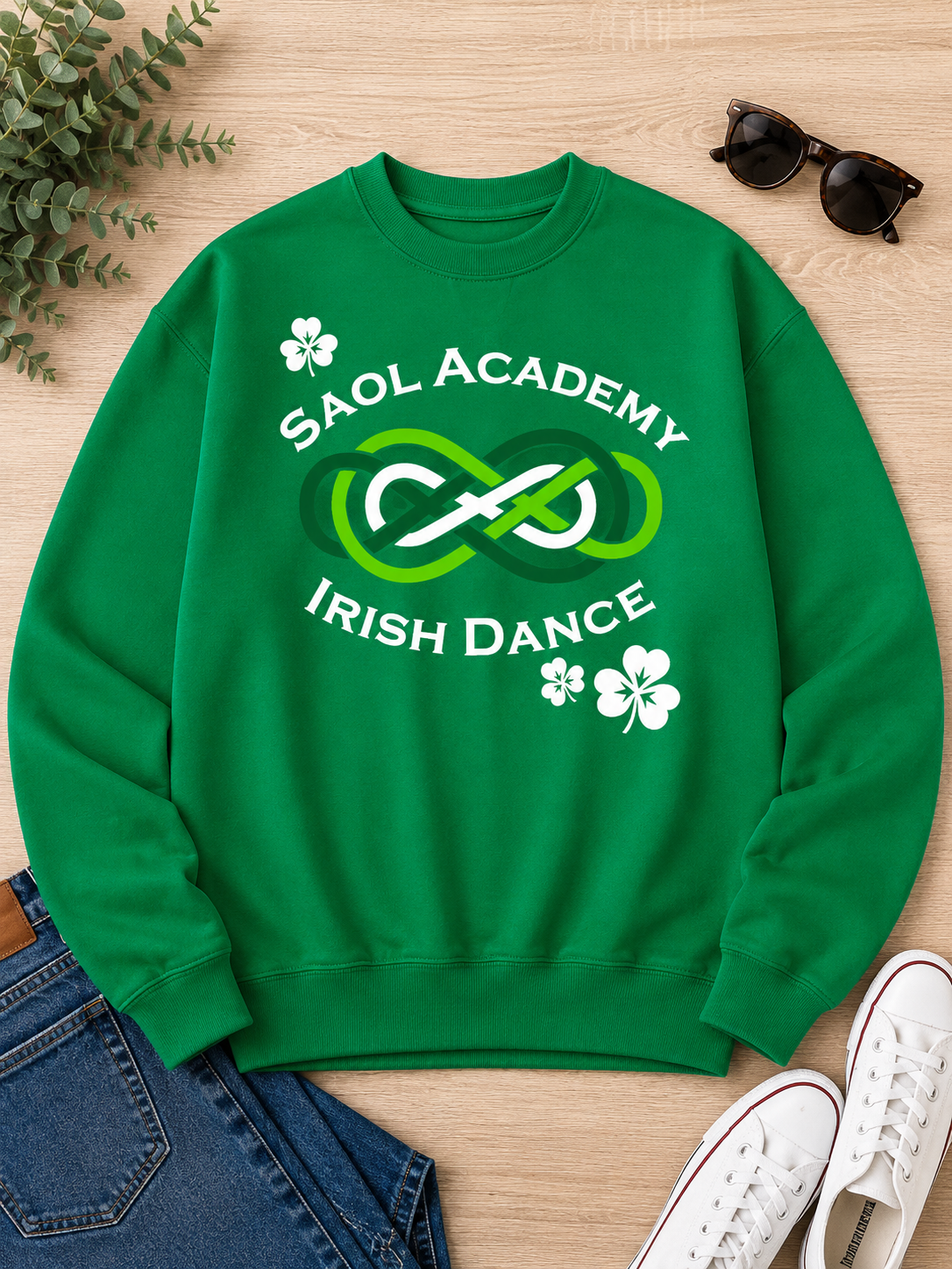 A green crewneck sweatshirt with a 'Saol Academy Irish Dance' graphic printed on the chest, styled in a flat lay with jeans, sunglasses, white sneakers, and a plant on a light wood background.