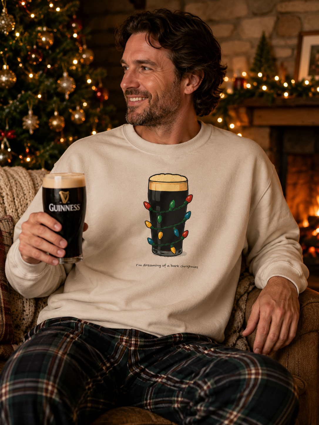 A man wearing a cream sweatshirt with a graphic of a pint of Guinness decorated with Christmas lights, sitting in a cozy living room with a fireplace and a Christmas tree in the background.