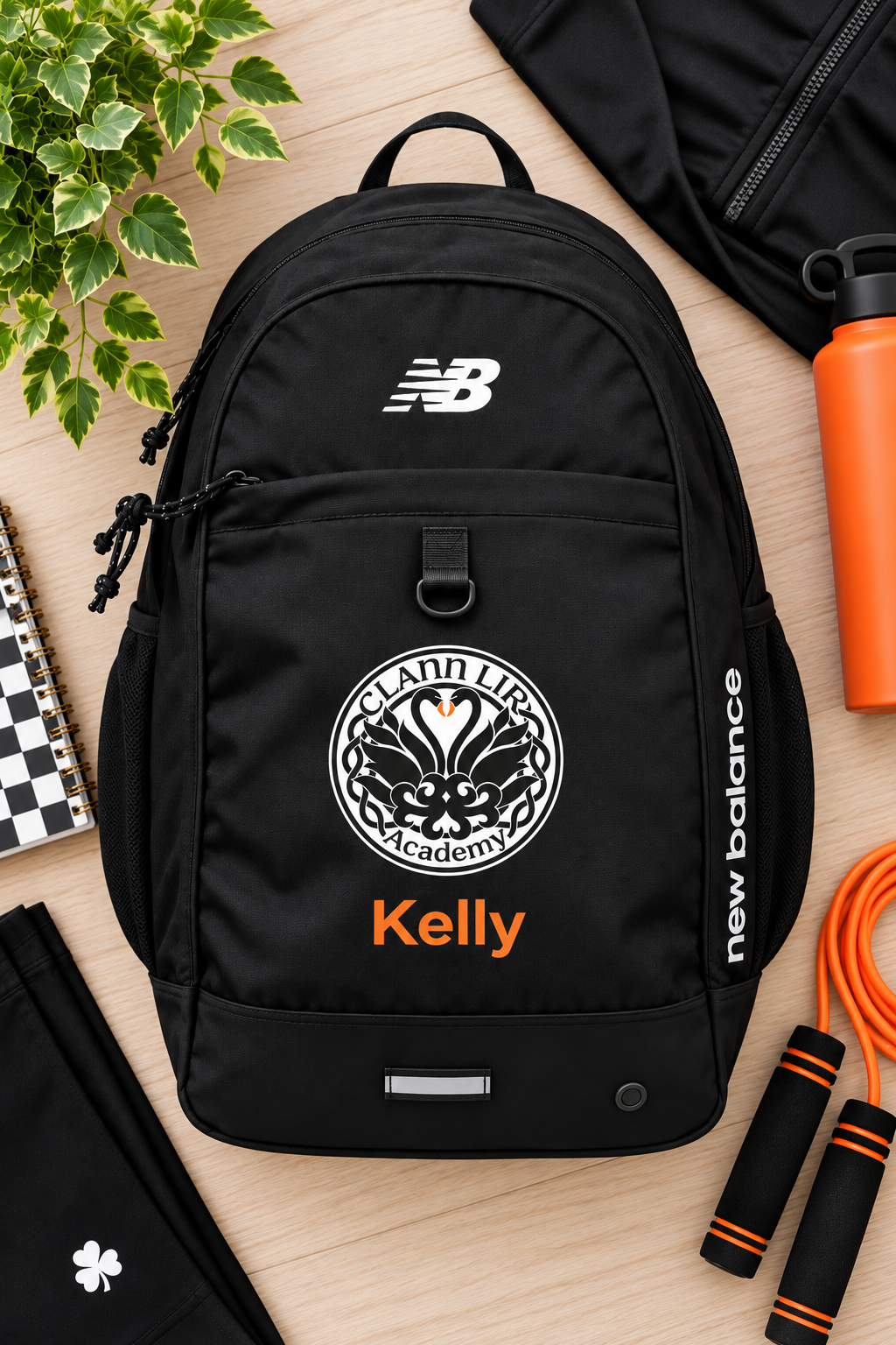 A black New Balance backpack laid flat on a light wood surface, decorated with a white circular Clann Lir Logo' in white and orange, and an orange 'new balance' logo on the side.