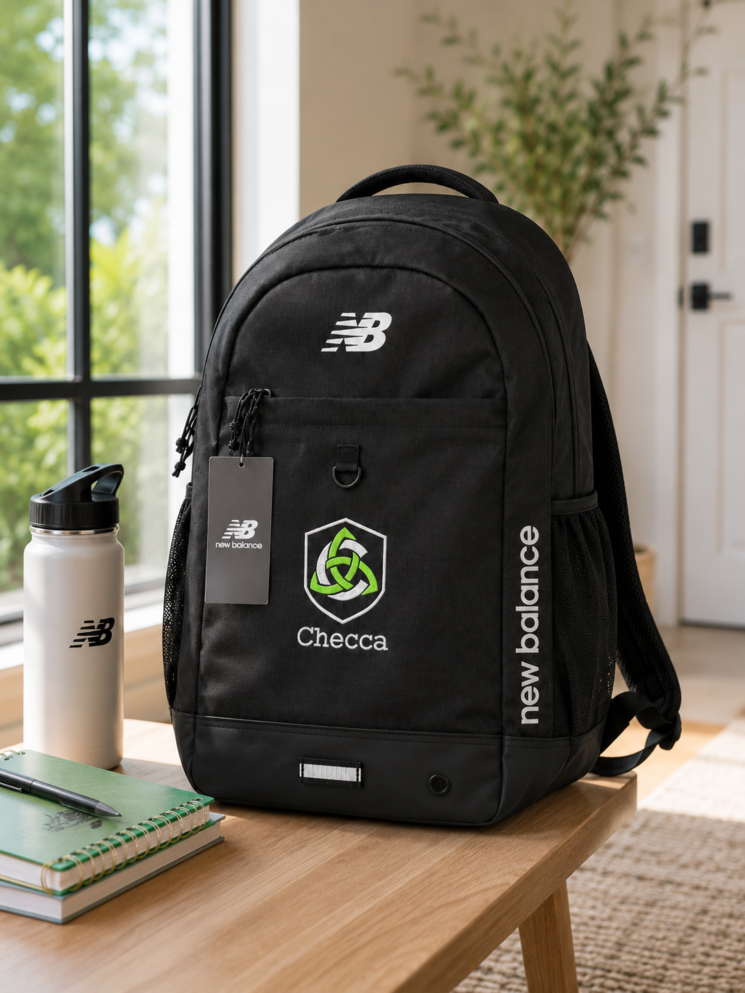 A black New Balance backpack with white and green logo graphics sits on a wooden table next to a water bottle and notebook in a bright, modern room.