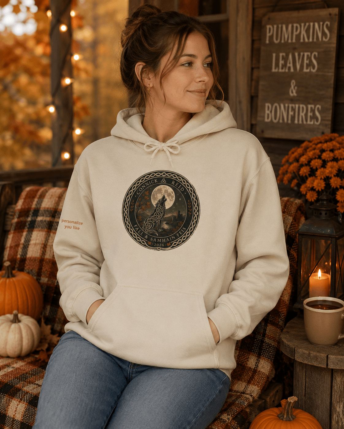 A woman wearing a cream-colored hoodie with a circular graphic of a bird in a tree and the words 'Winter Wonderland' sitting in a cozy, autumn-themed setting with pumpkins, a candle, and a wooden sign that reads 'Pumpkins Leaves & Bonfires'.
