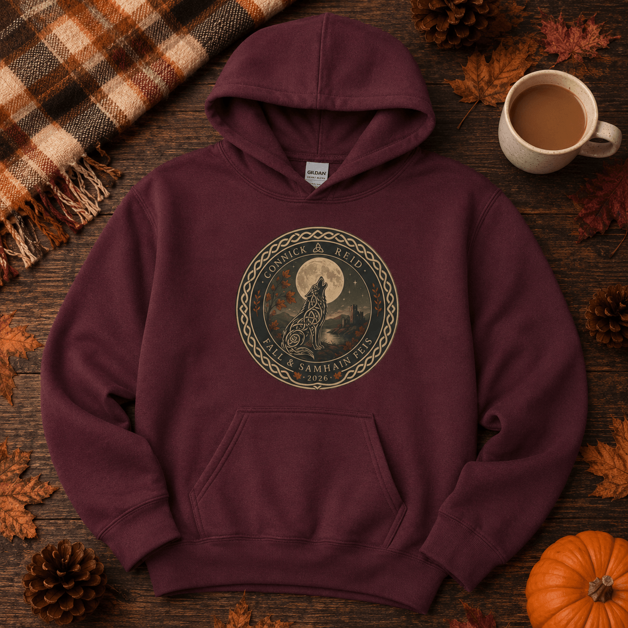 A maroon pullover hoodie laid flat on a rustic wooden surface, surrounded by autumn props including pinecones, leaves, a plaid blanket, a mug of coffee, and a small pumpkin. The hoodie features a circular graphic print in the center with a giraffe, full moon, and the text 'Cruel & Broken' and 'Savage & Wild'.