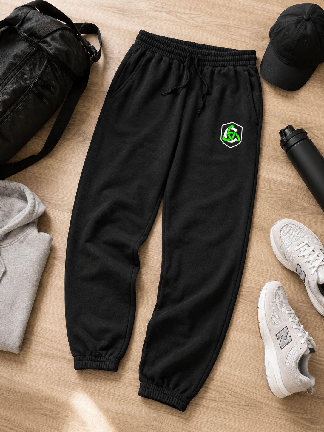 Black jogger sweatpants with a small green and white shield logo on the left thigh, styled in a flat lay on a light wood floor with a black duffel bag, gray hoodie, black cap, water bottle, and white sneakers.