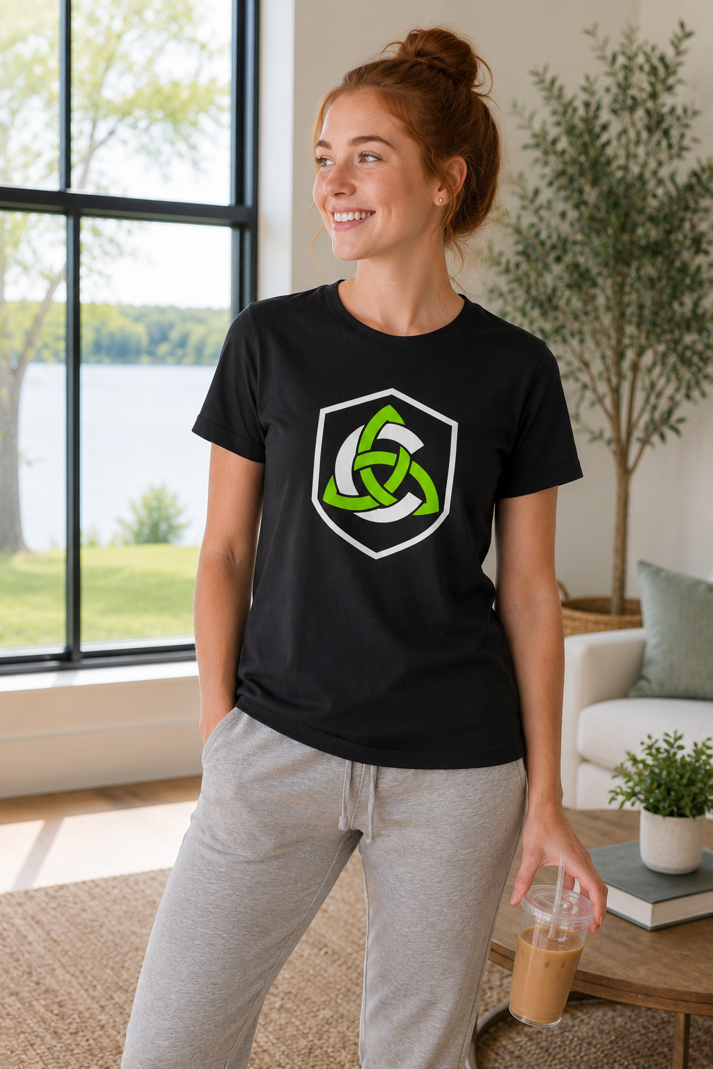 A woman wearing a black t-shirt with a green and white Celtic knot logo, paired with light gray sweatpants, standing in a bright living room.