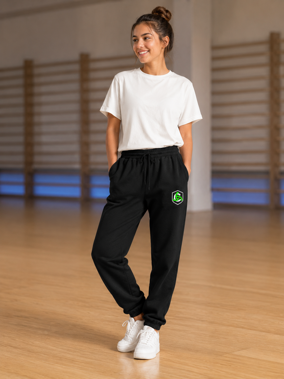 A woman standing in a gym wearing black sweatpants with a green shield logo on the left thigh, a white t-shirt, and white sneakers.