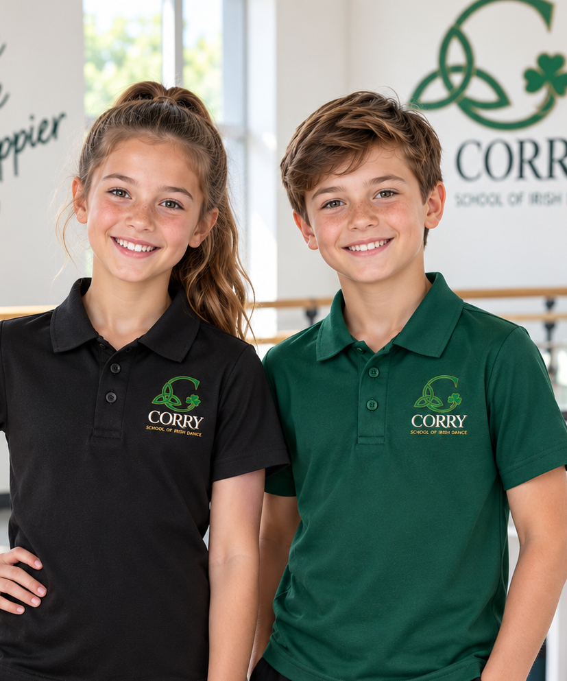 Two young dancers, a girl in a black polo shirt and a boy in a green polo shirt, both featuring the 'Corry School of Irish Dance' logo on the left chest, standing in a bright indoor studio environment.